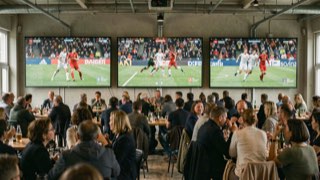 Drie schermen naast elkaar met drie verschillende Europese wedstrijden, publiek eronder aan hoge tafels.