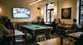 Brede opname van een cafézaal in avondlicht met een scherm, een biljart en een dartbord in één beeld.