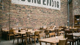 Breed scherm hoog aan een bakstenen muur, daaronder rijen tafels met stoelen schuin naar voren gedraaid.