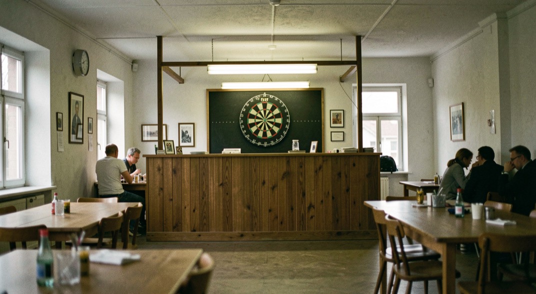 Hoek van een zaal met een dartbord onder eigen verlichting, afgeschermd van de tafels door een balie.